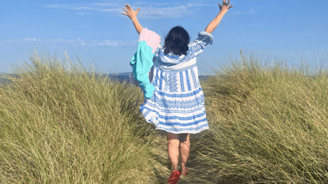 A woman walking through dune grass with her arms raised to a blue sky