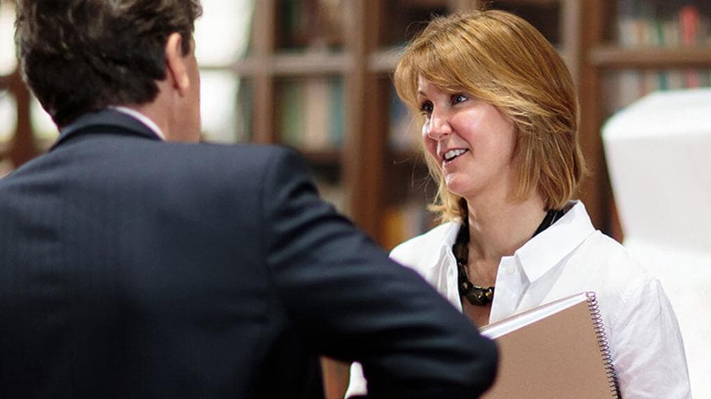 An alumna holding a notebook smiles as she talks with a colleague in a library