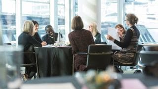 Participants in conversation around a table in a bright room with tall windows