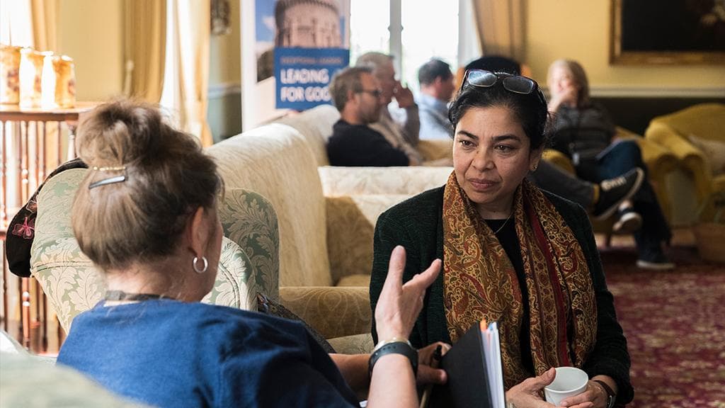 Two participants in discussion in a lounge, other attendees talking in the background